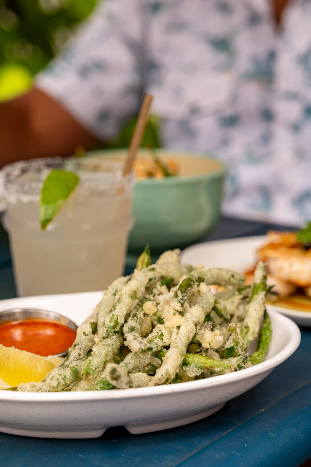 a plate of fried green vegetables with a small bowl of red sauce and a lemon wedge on a blue wooden table