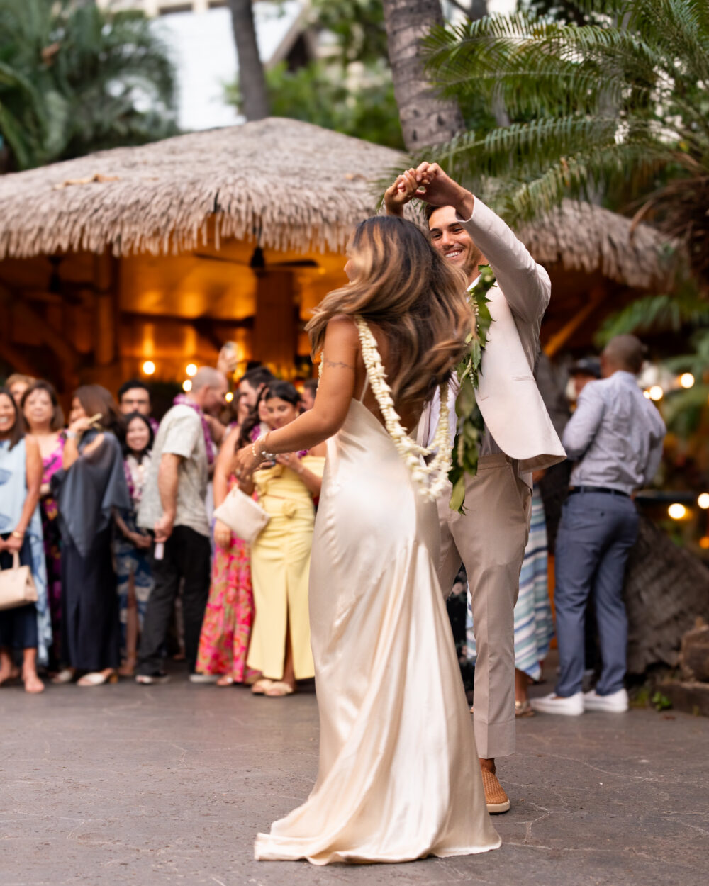 A couple dances in front of a thatched roof tiki hut with a crowd of people.