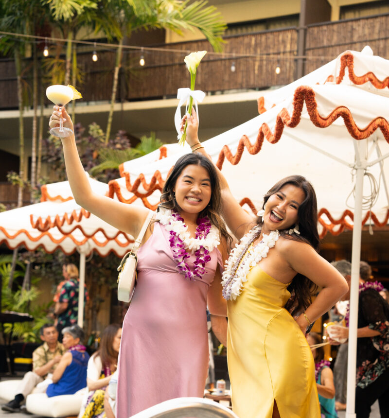 Two women in leis posing with drinks and a flower