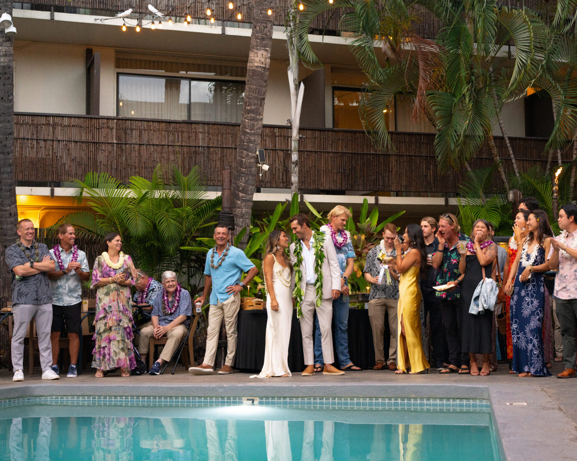 A group of people in formal and casual attire standing by a pool with a building in the background