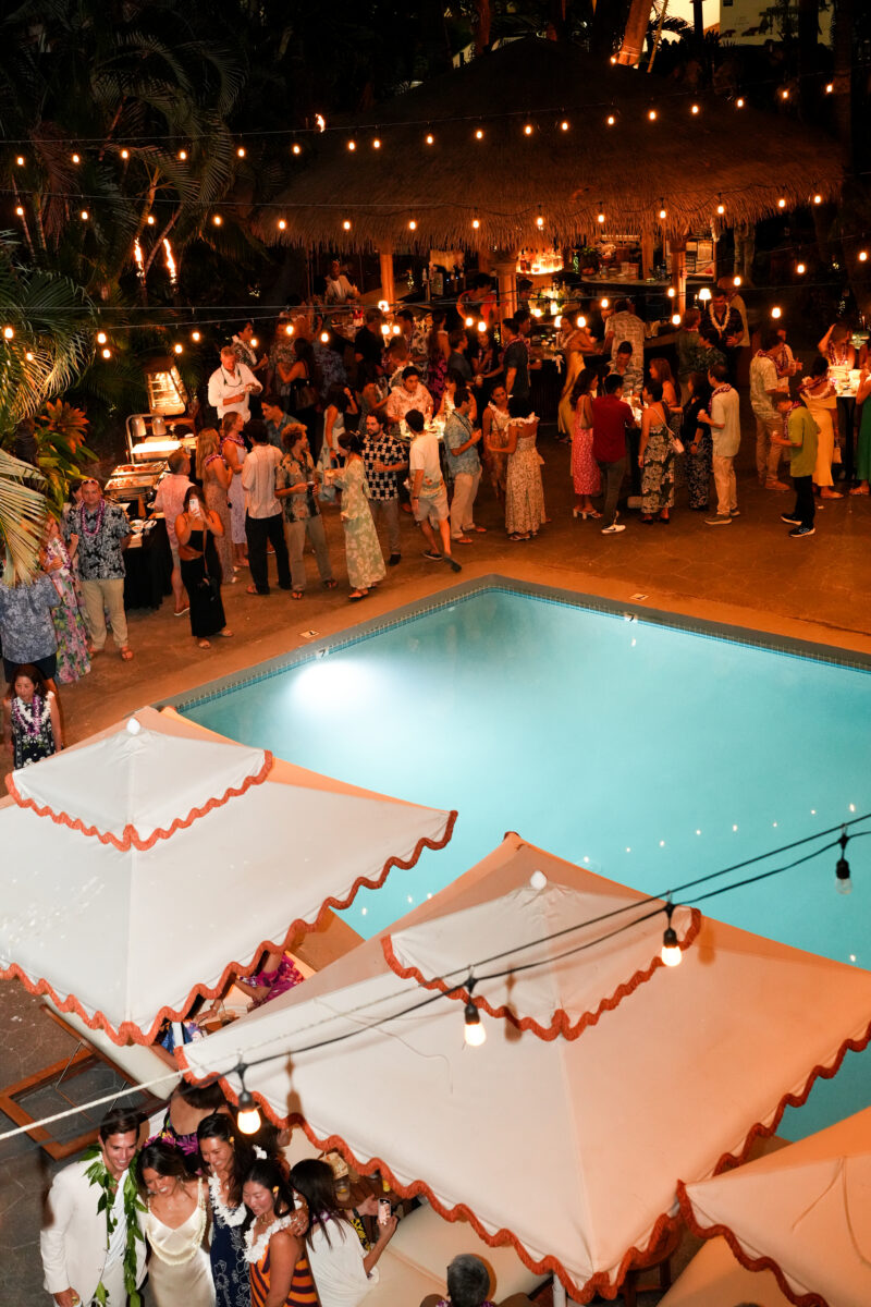 A nighttime view of a pool party with people gathered around the pool and string lights above.